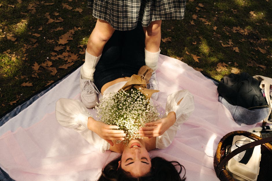 A young woman with long dark hair, lying on her back on a pink blanket spread over grass sprinkled with fallen leaves in a park, is smiling and holding a bouquet of small white baby's breath flowers w