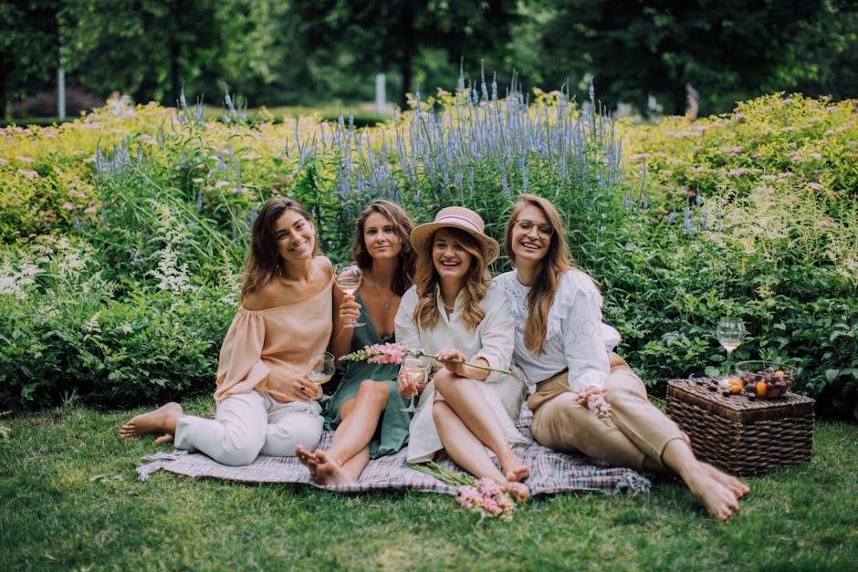 Four women sitting on a picnic blanket in a lush garden with blooming flowers and greenery, enjoying a floral picnic with glasses of wine. One woman wears a wide-brimmed straw hat and a white dress, h
