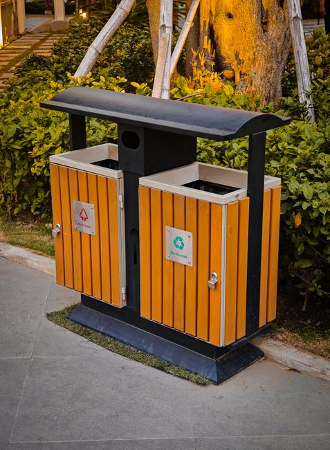 A pair of wooden recycling and waste bins situated on a paved sidewalk in an outdoor urban setting, with lush green shrubbery and a large tree with textured bark and yellow foliage in the background.
