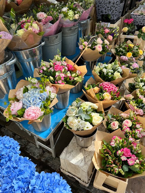 A diverse selection of fresh flower bouquets arranged in metal buckets and wrapped in brown craft paper, featuring roses, hydrangeas, and carnations in shades of pink, white, and blue. The flowers are