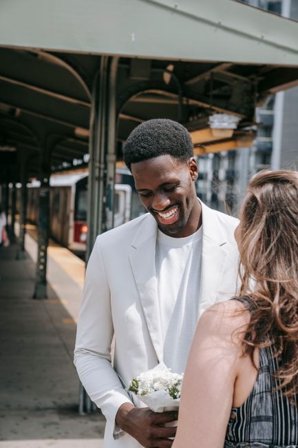 A smiling man of African descent dressed in a white blazer holding a small bouquet of white and green flowers, engaging in a conversation with a woman whose back is to the camera. They are standing ou
